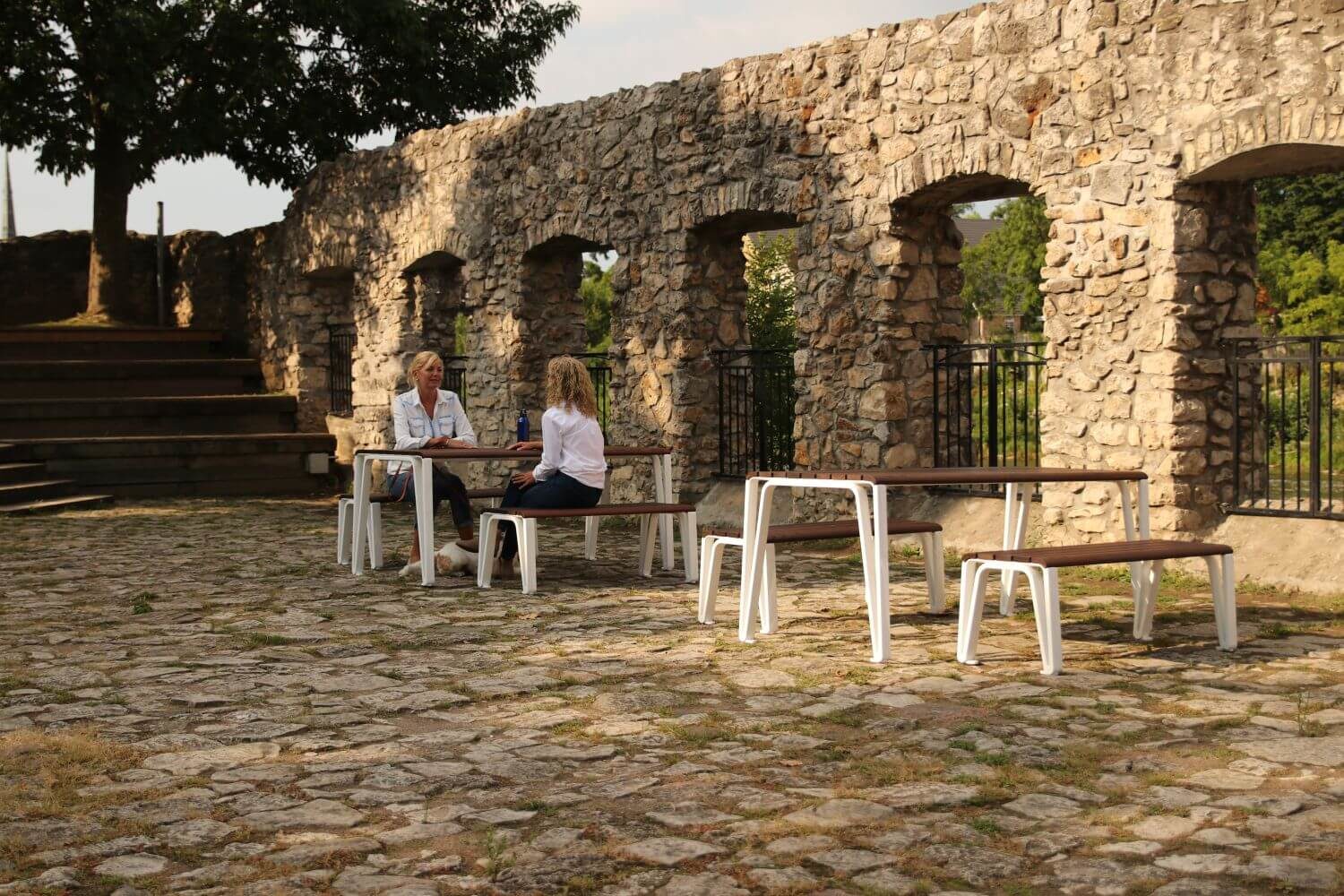 HOME_INSTALLATIONS_TABLES & SEATING_Maglin Photoshoot_Iconic Table & Benches white legged tables with wood top. In the background two women are sitting at a table
