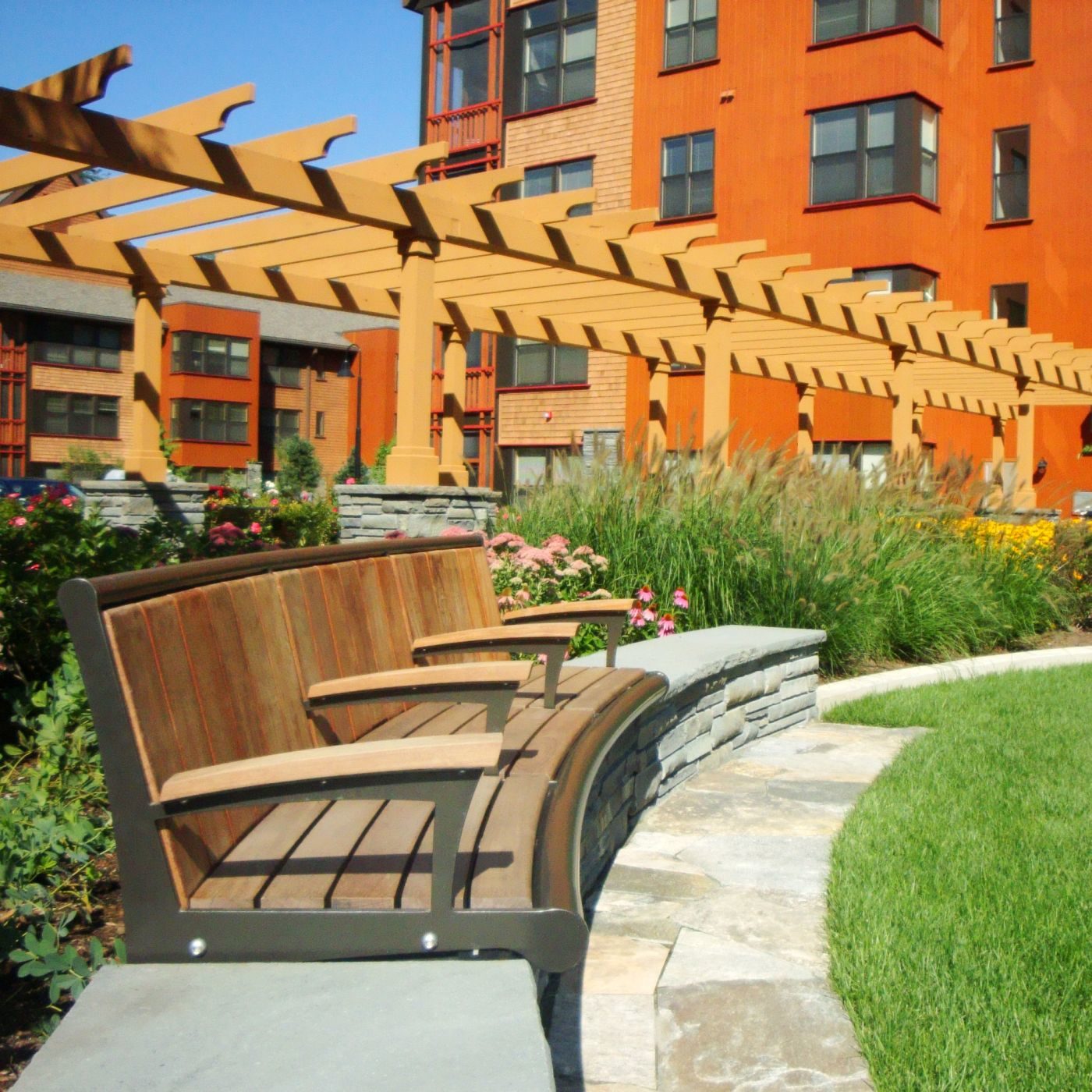 Banc en bois sur un patio en pierre devant des jardins paysagers, avec une pergola en bois et un bâtiment orange en arrière-plan sous un ciel bleu clair.