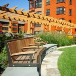 Banc en bois sur un patio en pierre devant des jardins paysagers, avec une pergola en bois et un bâtiment orange en arrière-plan sous un ciel bleu clair.