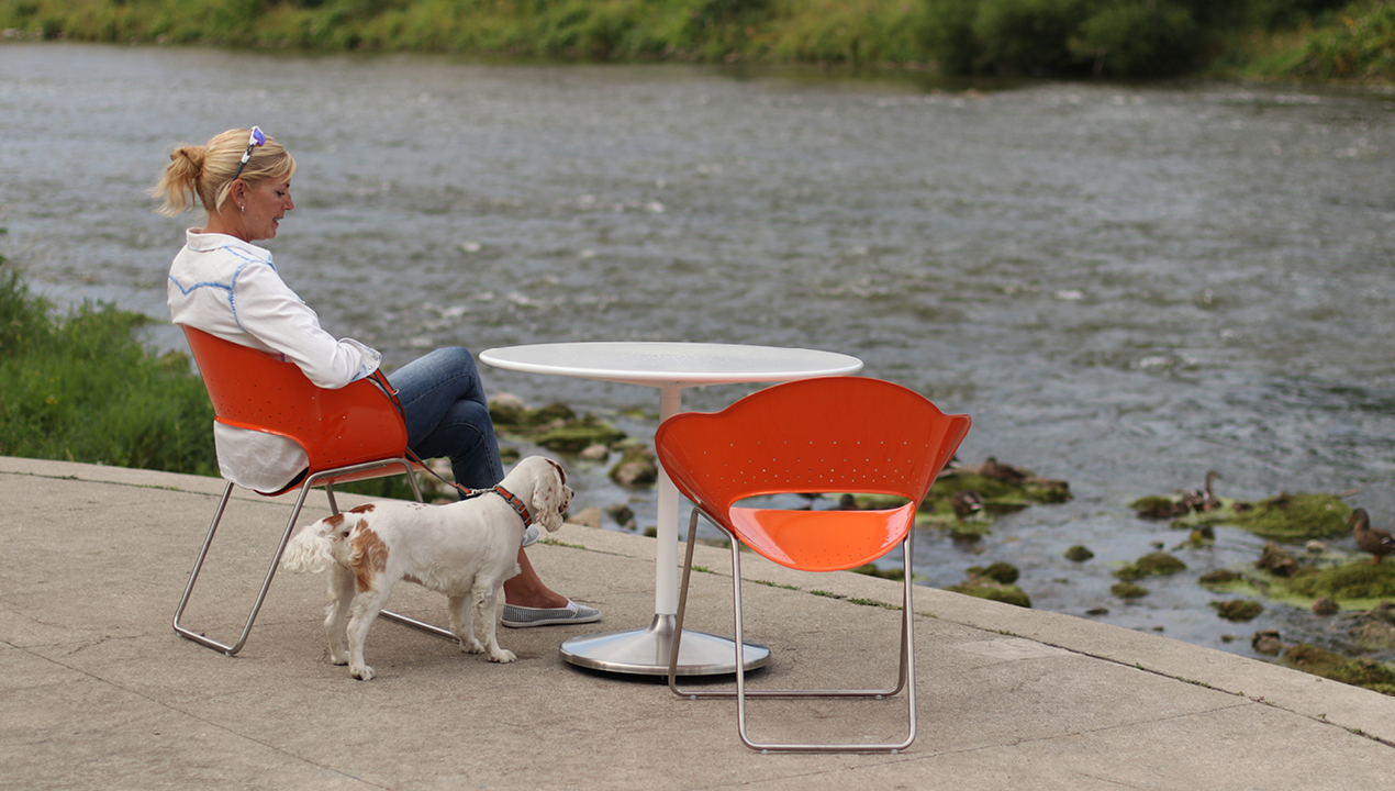Woman and Dog sitting on Orange Battery Collection Chairs facing the river