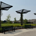 Three metal shade structures with hexagonal patterns stand along a pathway with benches, in a suburban park with grass, trees, and houses in the background.