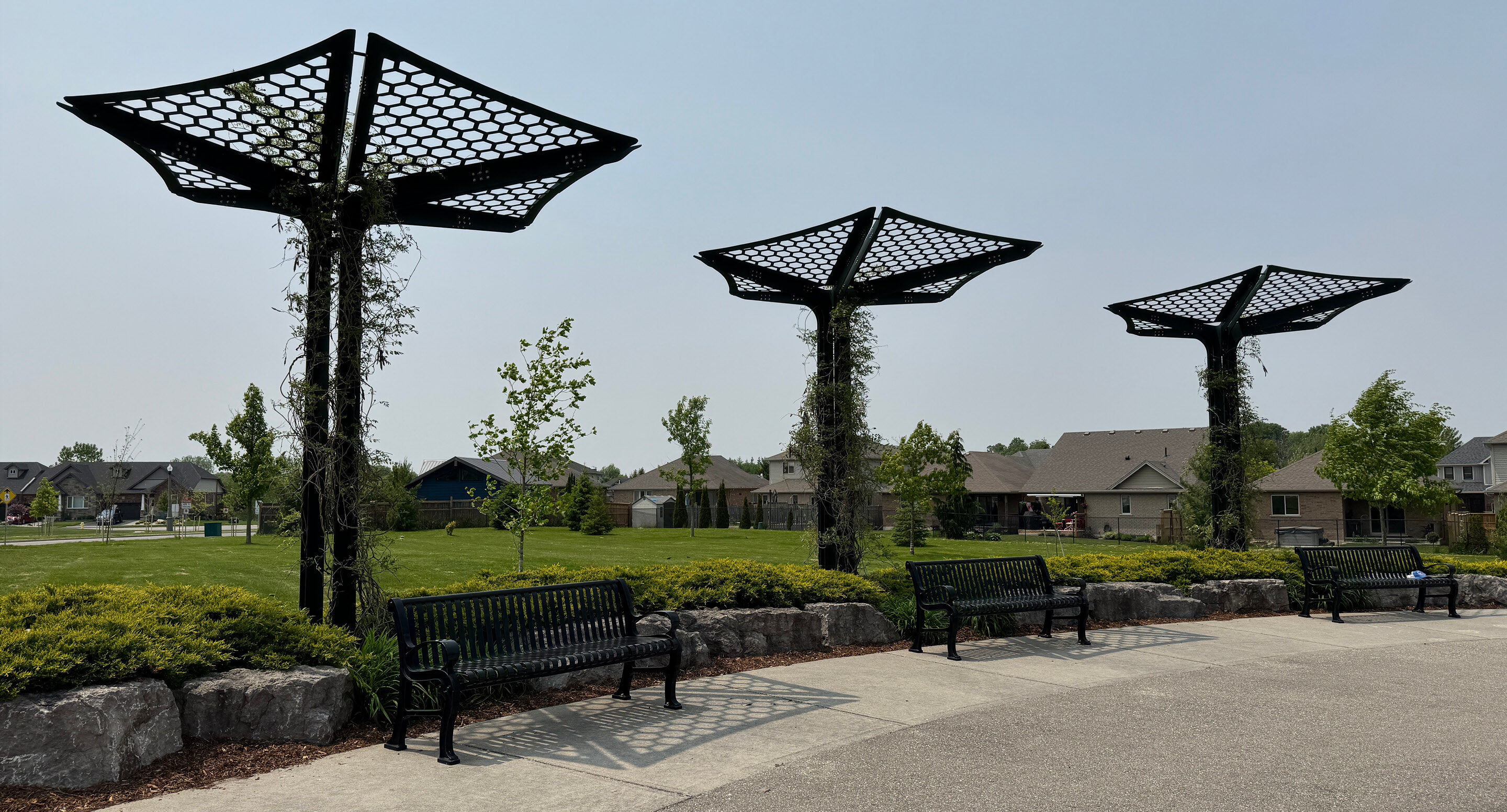 Three metal shade structures with hexagonal patterns stand along a pathway with benches, in a suburban park with grass, trees, and houses in the background.