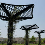 Trois structures métalliques noires à mailles hexagonales, ressemblant à des arbres abstraits, se dressent dans un parc herbeux avec des maisons et des arbres en arrière-plan. Des plantes grimpantes grimpent sur ces éléments d'infrastructure ombragés, mêlant ainsi la nature au design.