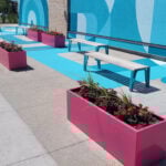 Outdoor seating area with white benches and pink rectangular 1500 planters filled with flowers, offering inspiration beside a blue patterned wall—an inviting installation set along the sidewalk.