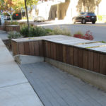 Wood and concrete benches line a sidewalk near a street with cars; plants and pedestrians are visible in the background.