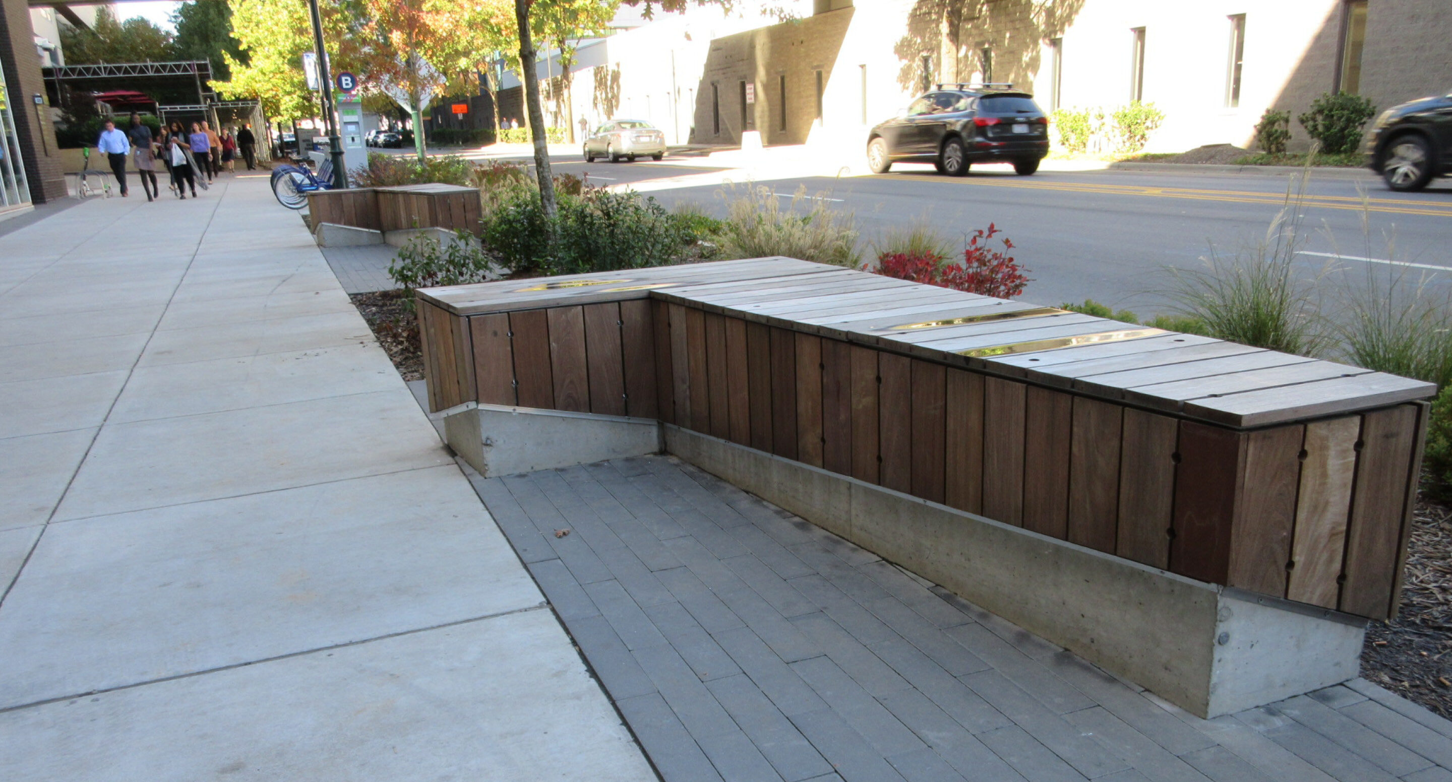 Wood and concrete benches line a sidewalk near a street with cars; plants and pedestrians are visible in the background.
