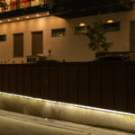 A wooden planter box with built-in lighting sits on a city sidewalk at night, with cars and a modern building in the background.