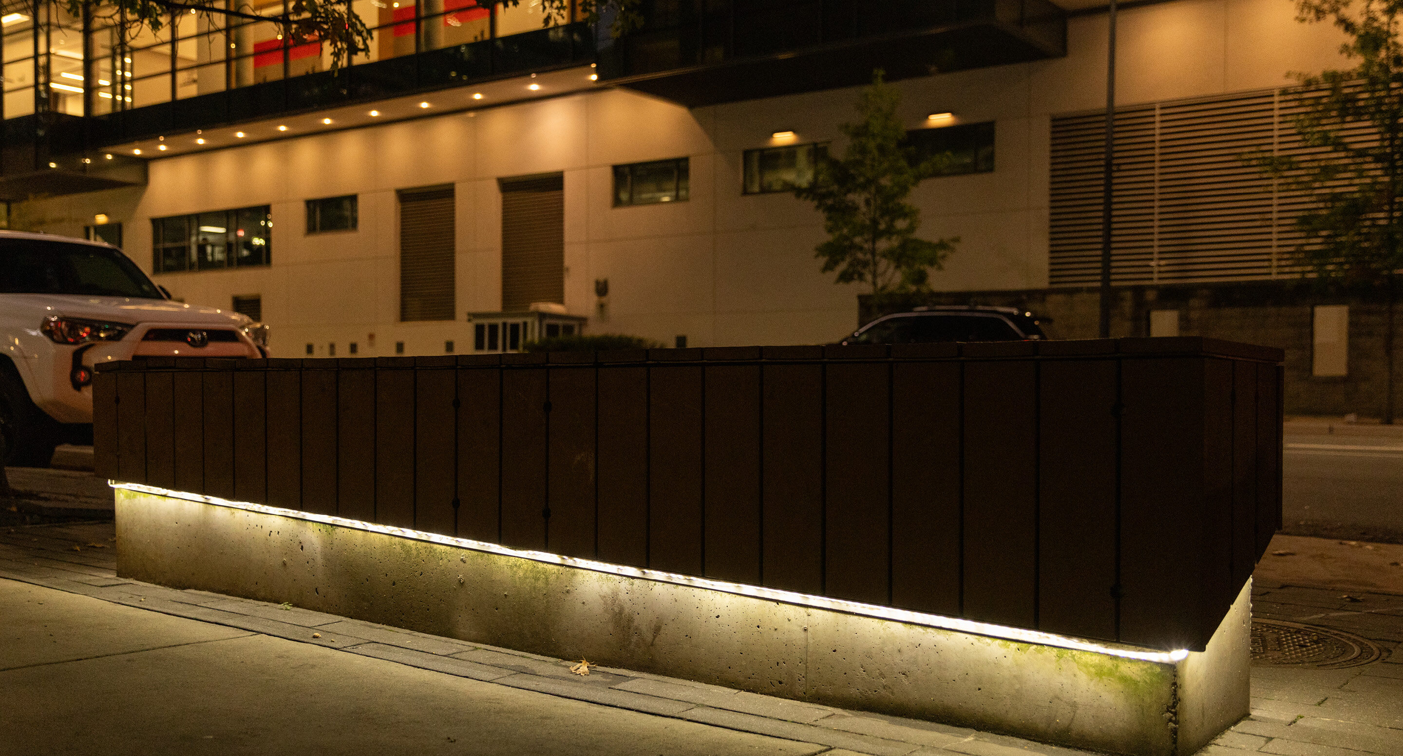 A wooden planter box with built-in lighting sits on a city sidewalk at night, with cars and a modern building in the background.