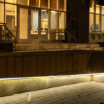 A wooden L-shaped bench with base lighting stands outside a building with glass doors at night; a person walks in the background.