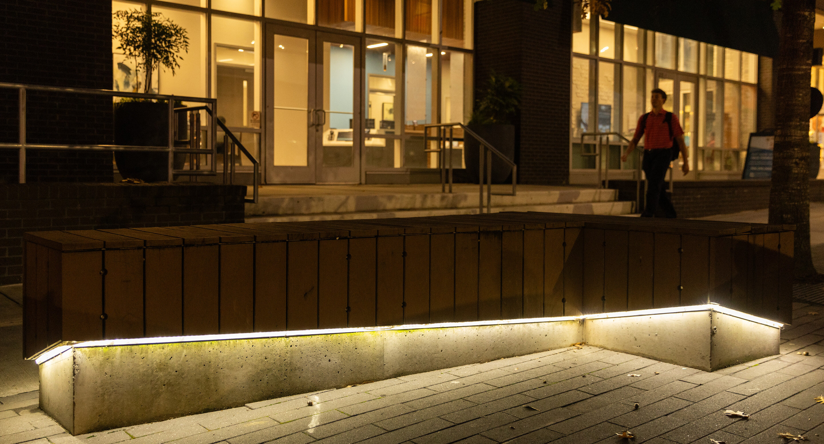 A wooden L-shaped bench with base lighting stands outside a building with glass doors at night; a person walks in the background.