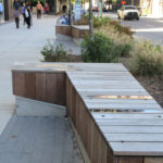 Wooden bench with metal accents on a city sidewalk, adjacent to landscaped plants and a street with cars and pedestrians in the background.