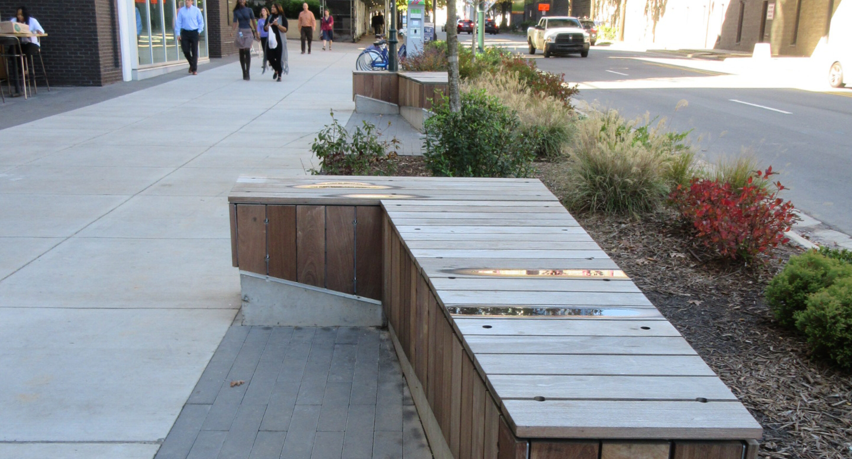 Wooden bench with metal accents on a city sidewalk, adjacent to landscaped plants and a street with cars and pedestrians in the background.