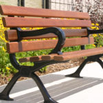 A 100 Backed Benches brown wooden bench with black metal armrests and legs sits on a concrete path by a flower bed filled with yellow flowers and green plants.