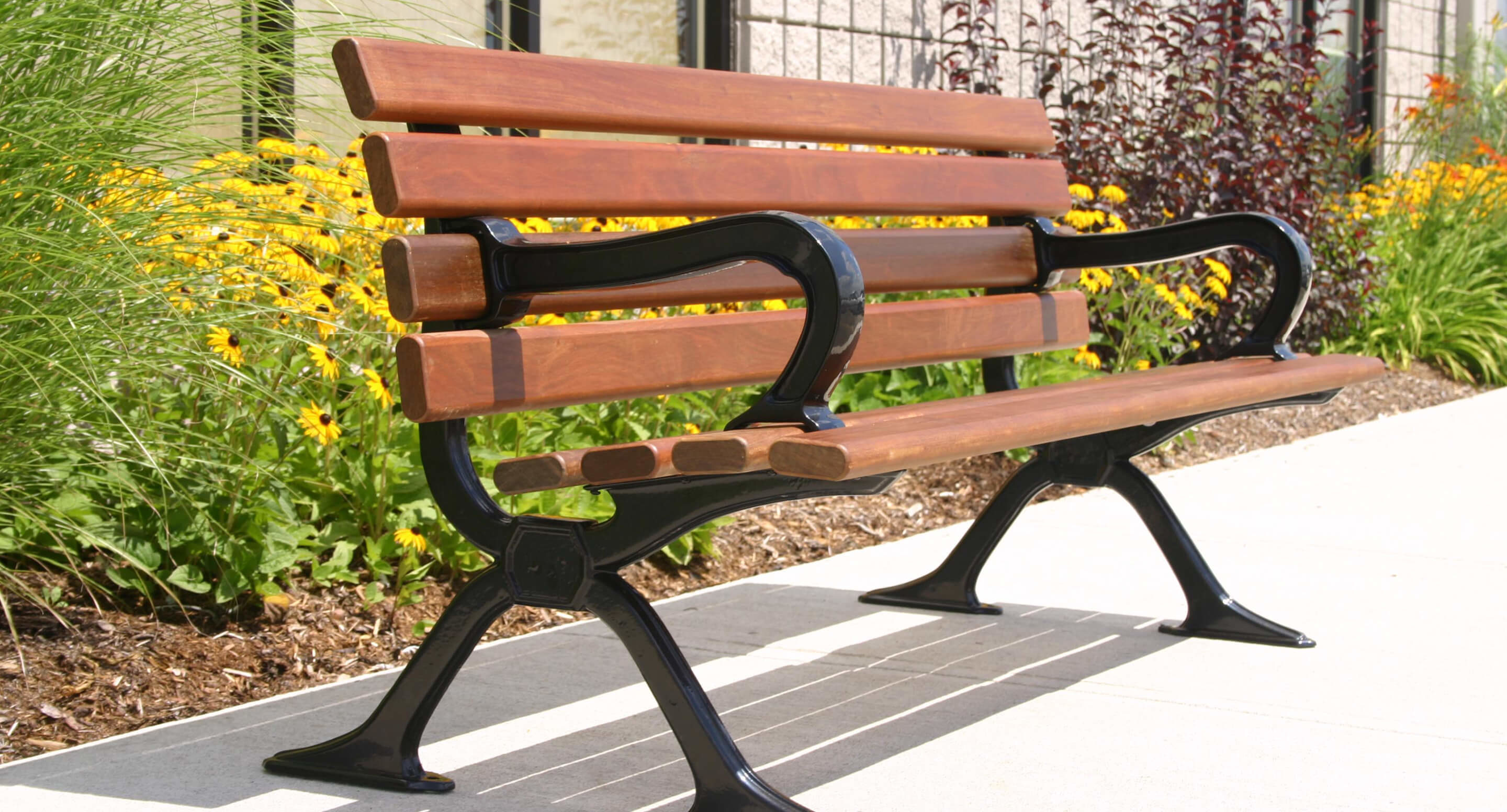 A 100 Backed Benches brown wooden bench with black metal armrests and legs sits on a concrete path by a flower bed filled with yellow flowers and green plants.