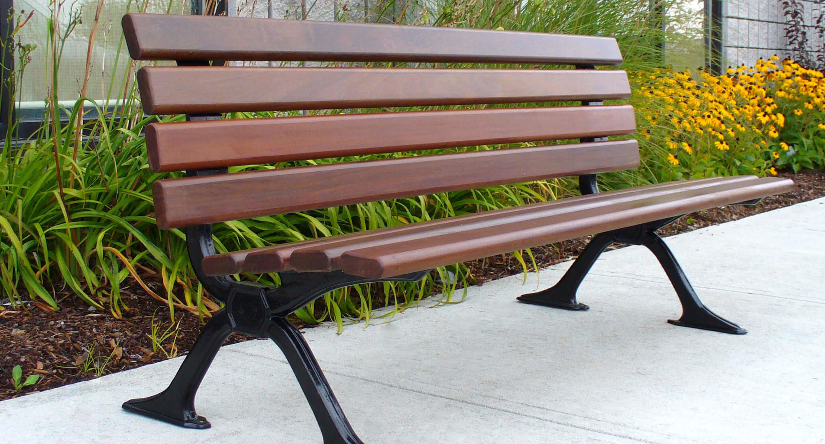 A 100 Backed Benches wooden park bench with black metal legs is placed on a concrete path beside green plants and yellow flowers.