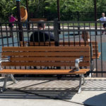 A 100 Backed Bench faces a fenced playground where children play in the background; a trash can is visible on the right side.