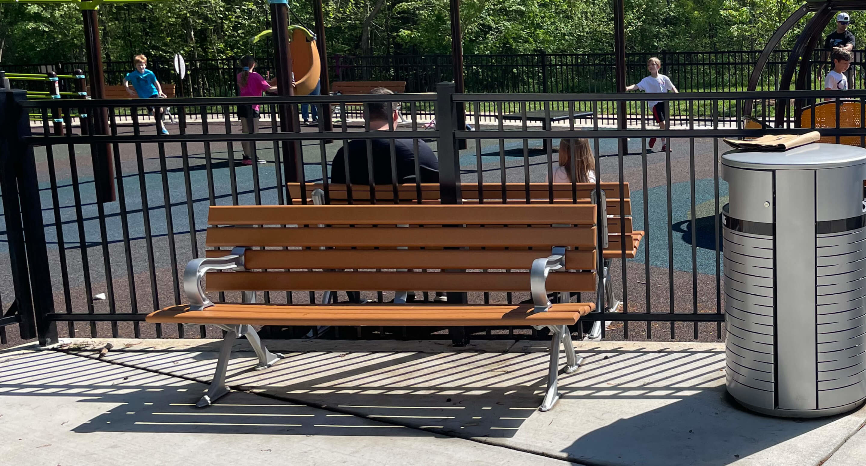 Le Banc avec dossier 100 fait face à une aire de jeux clôturée avec des enfants et des adultes, une poubelle sur la droite et des arbres en arrière-plan.