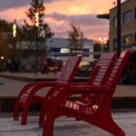 Three red outdoor chairs are aligned on a paved surface with a blurred background of buildings, trees, and a sunset sky.