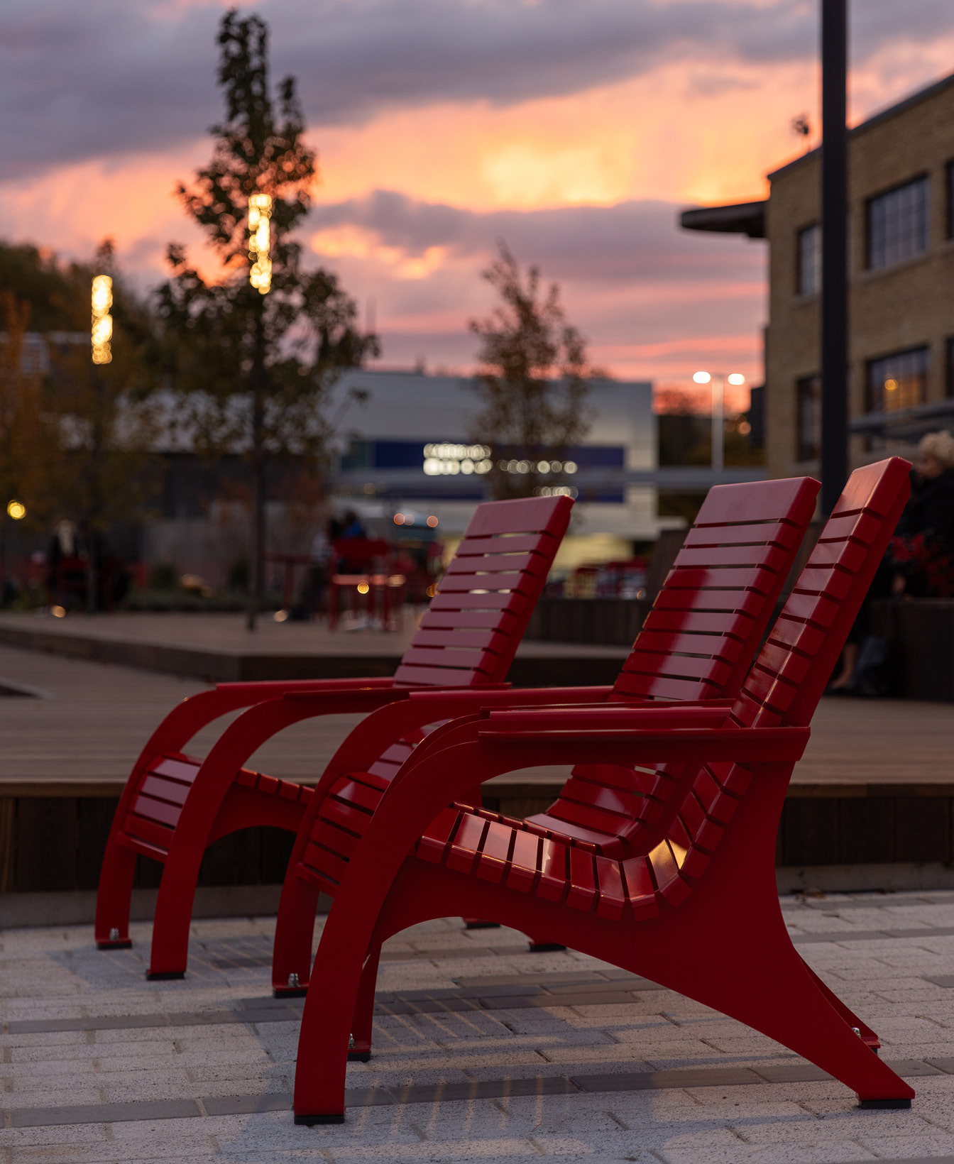 Three red outdoor chairs are aligned on a paved surface with a blurred background of buildings, trees, and a sunset sky.