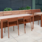 Two wooden tables with matching Foro Café Chairs are set on a tiled patio beside a long wooden bench, lush plants, and a building featuring large windows and black blinds.