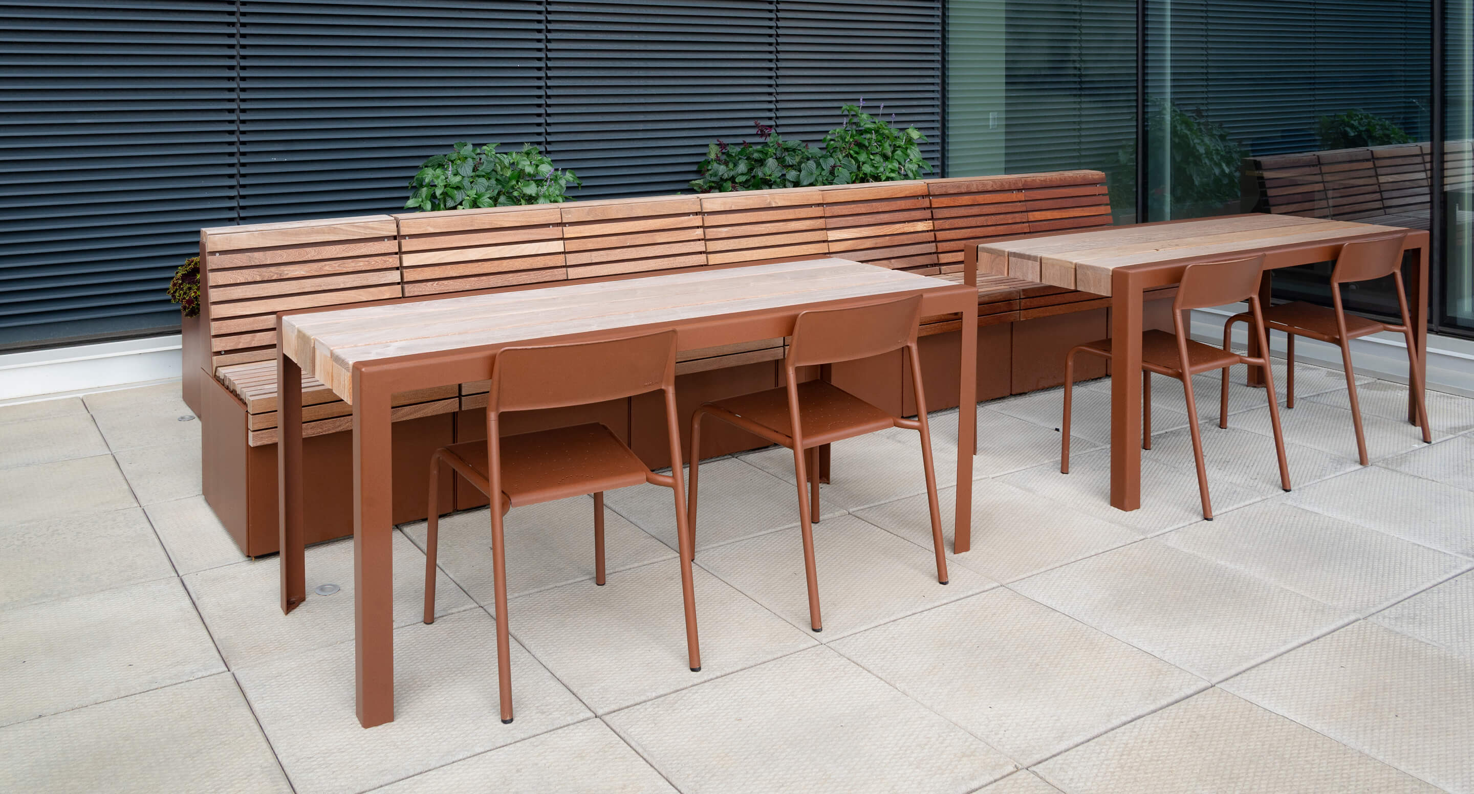 maglin-pixel-1050-foro_1 Two wooden tables with matching Foro Café Chairs are set on a tiled patio beside a long wooden bench, lush plants, and a building featuring large windows and black blinds.