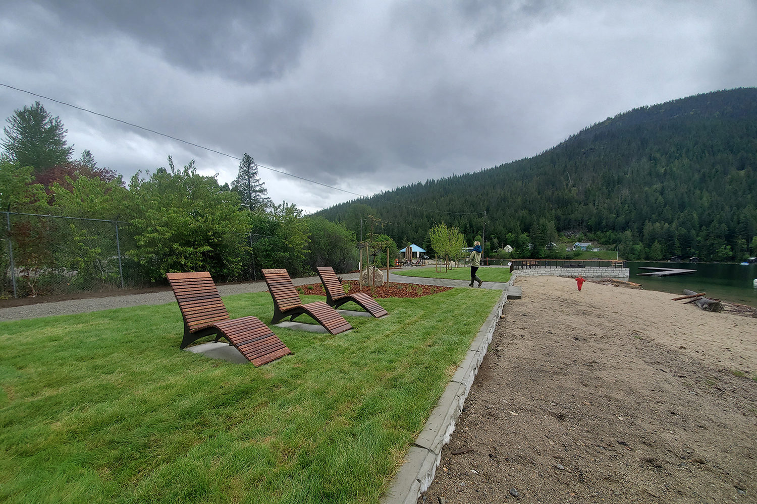 Three wooden lounge chairs on grass facing a lakefront with forested mountains in the background