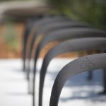 Close-up of metal bike racks in a row on a sidewalk, each featuring a bicycle symbol, with a blurred background of greenery and mulch.