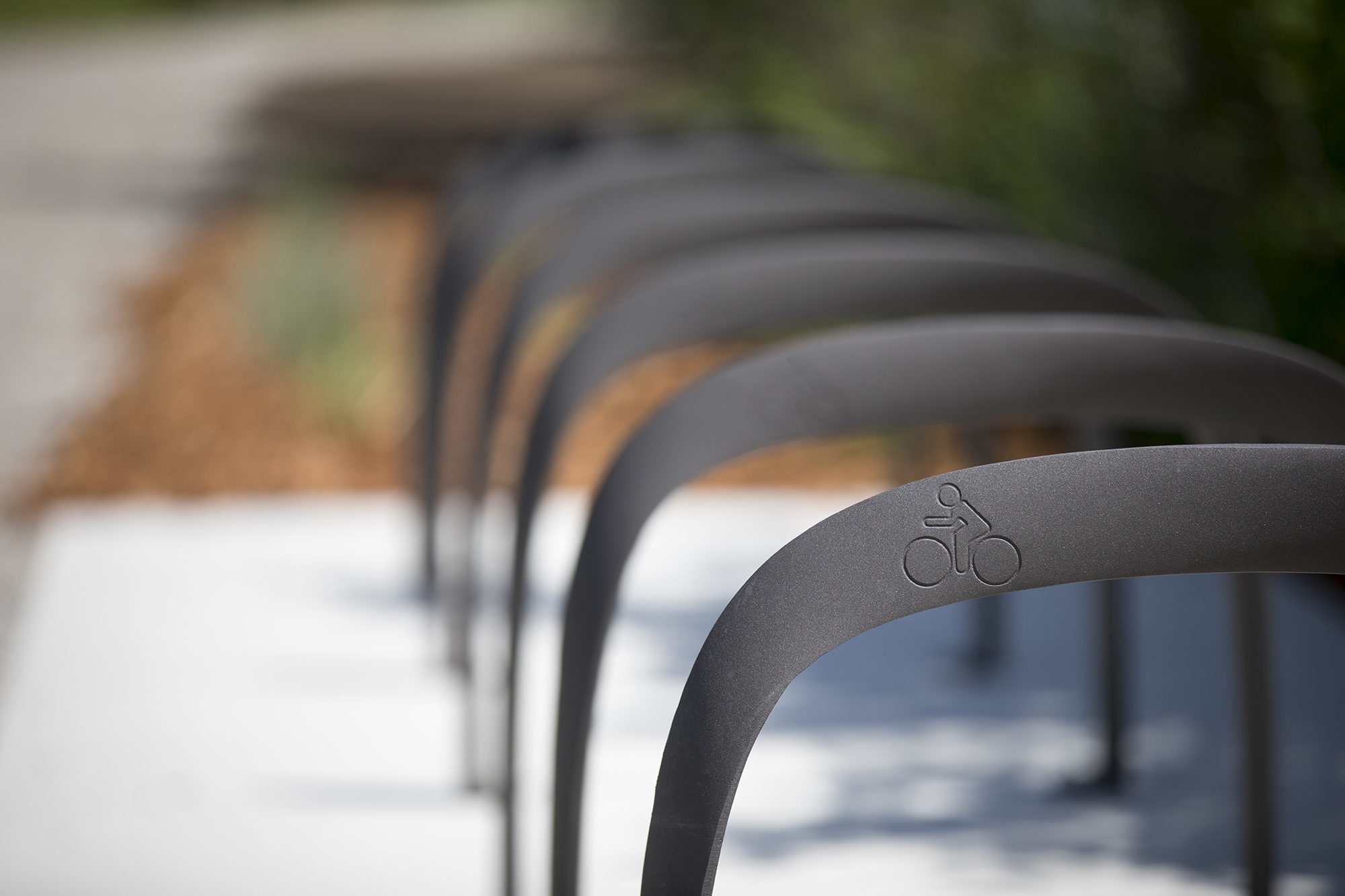 Close-up of metal bike racks in a row on a sidewalk, each featuring a bicycle symbol, with a blurred background of greenery and mulch.