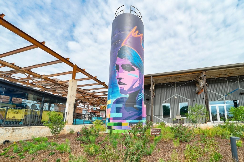 A water tower is wrapped in a mural featuring a woman in the foreground. In the background is an old warehouse facility turned into office space and dining