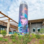 A water tower is wrapped in a mural featuring a woman in the foreground. In the background is an old warehouse facility turned into office space and dining