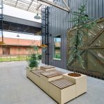 A modern outdoor courtyard features wooden benches with built-in planters, surrounded by metal walls and large rustic doors, under a partially covered roof.