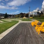 Five yellow lounge chairs are lined up on a wooden deck at the top of a terraced grass park. More yellow lounge chairs are in the background