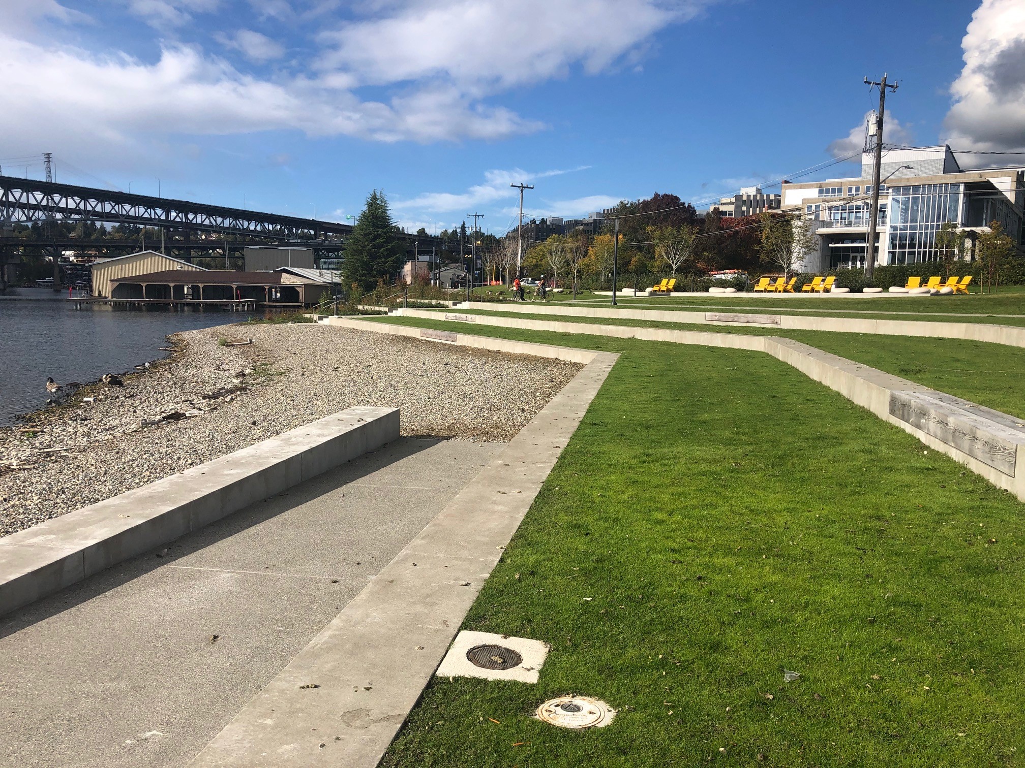 Foreground shows terraced lawn leading to a pebble beach. In the background, at the top of the terraced lawn, there are several yellow lounge chairs.