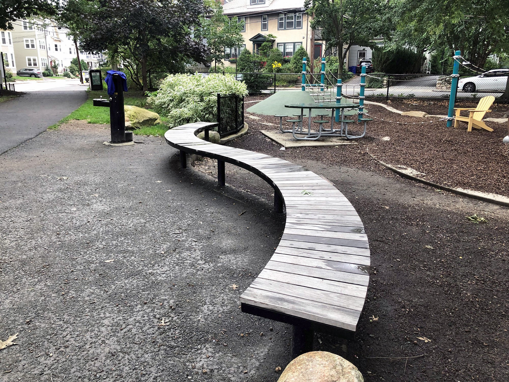 MA_Emerson Garden_Ogden_curved_backless_pedestal_Aged Ipe_001 A curved wooden Ogden bench is to the left of a small playground.
