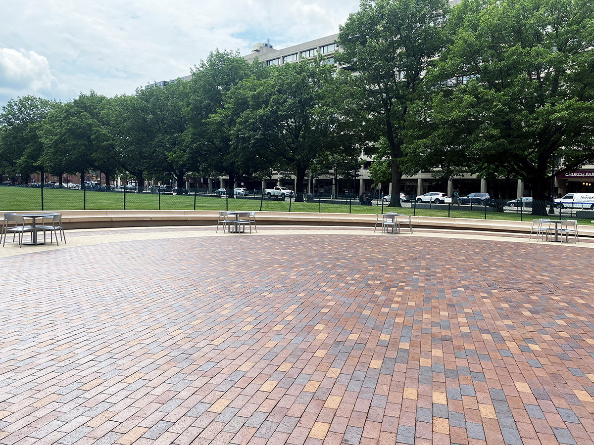 Image shows four sets of foro tables and chairs on a paved plaza. Green grass and trees are in the background.