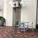 A Foro table and chairs on a paved plaza set before a concrete building. A planter is beside the table and large, round outdoor lighting fixtures are attached to the concrete building above the table.