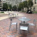 Two sets of Foro tables and chairs are shown on a paved plaza in the foreground. A busy city street is in the background.