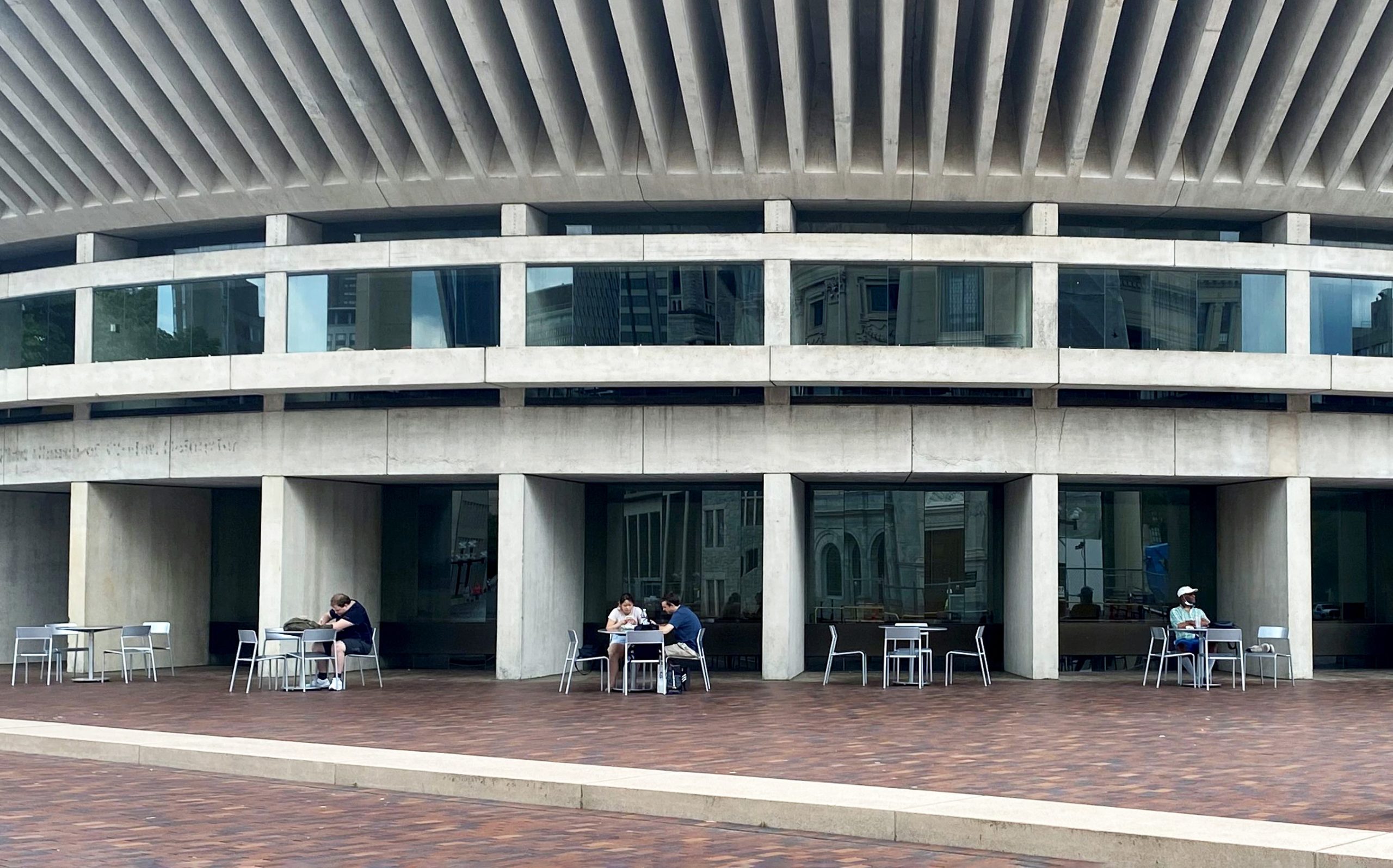 From a distance, people are seen sitting and eating in a plaza in front of a concrete building.