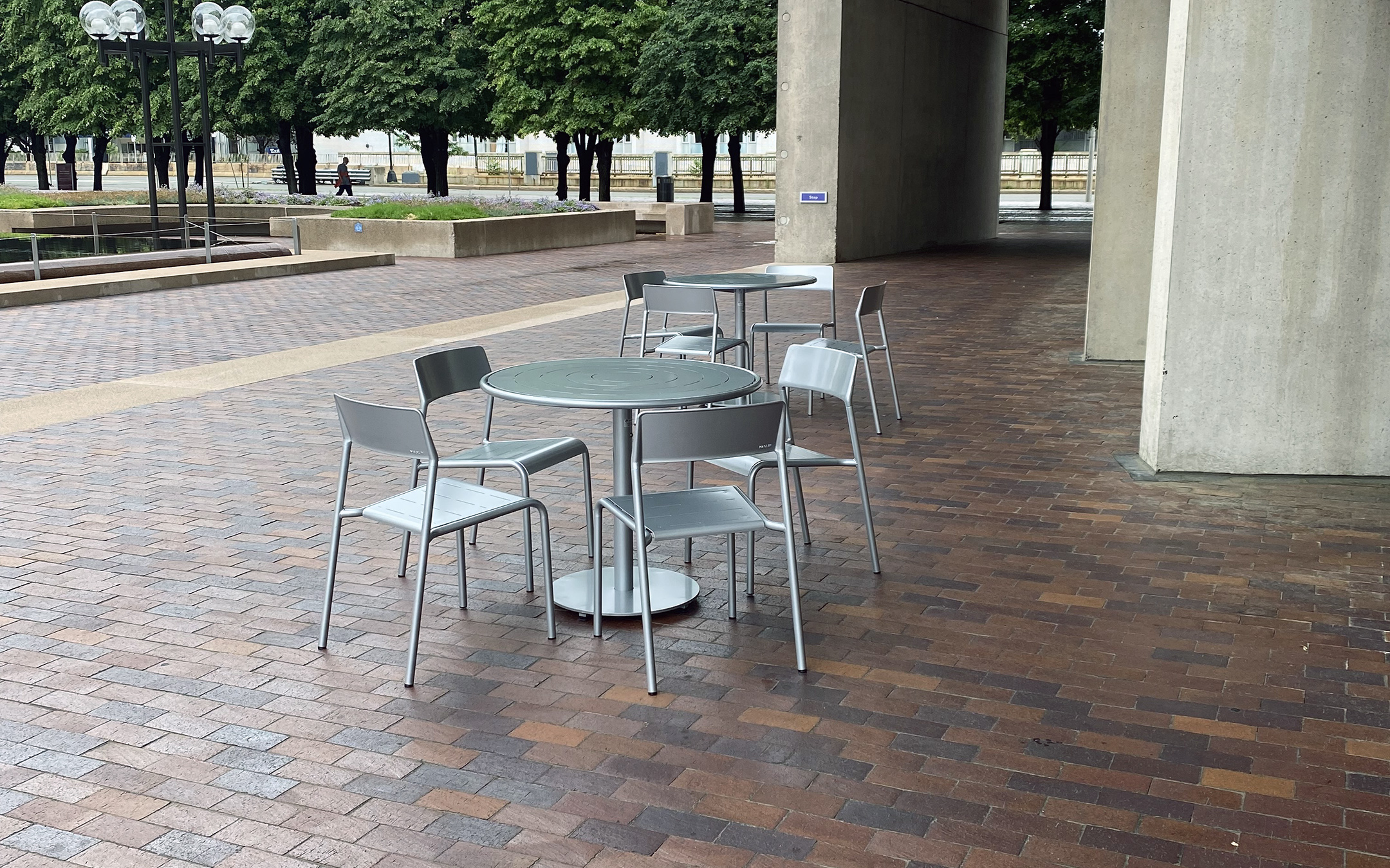 Two sets of ALUM Tables and Chairs are on a paved plaza next to a concrete building