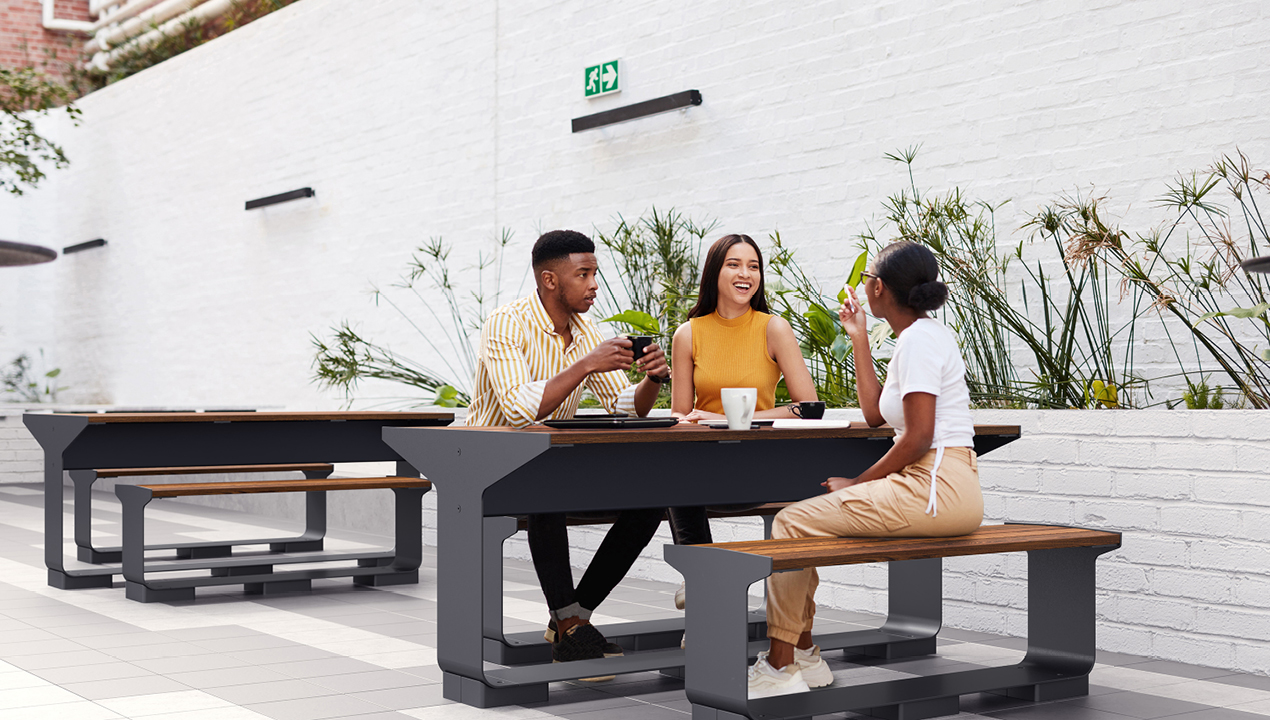 Three people sit at a picnic table outdoors, talking and drinking coffee against a white brick wall with plants in the background.