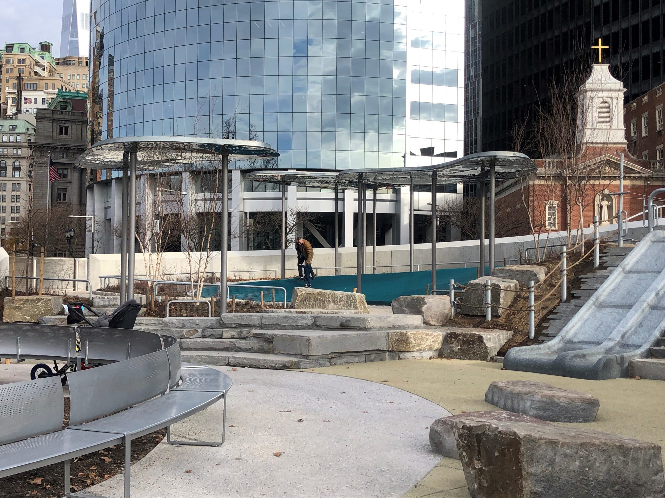 View of Battery Playscape with steel cloud canopies and curved bench. NYC buildings in the background