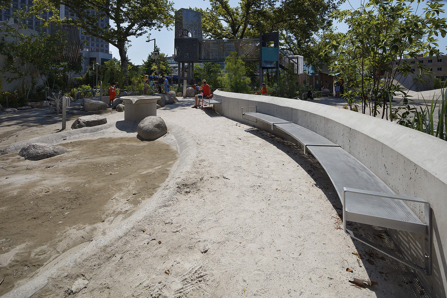 A curved concrete bench and sand area with scattered rocks in an outdoor playground, with children and adults in the background.