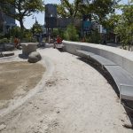 A curved concrete bench and sand area with scattered rocks in an outdoor playground, with children and adults in the background.