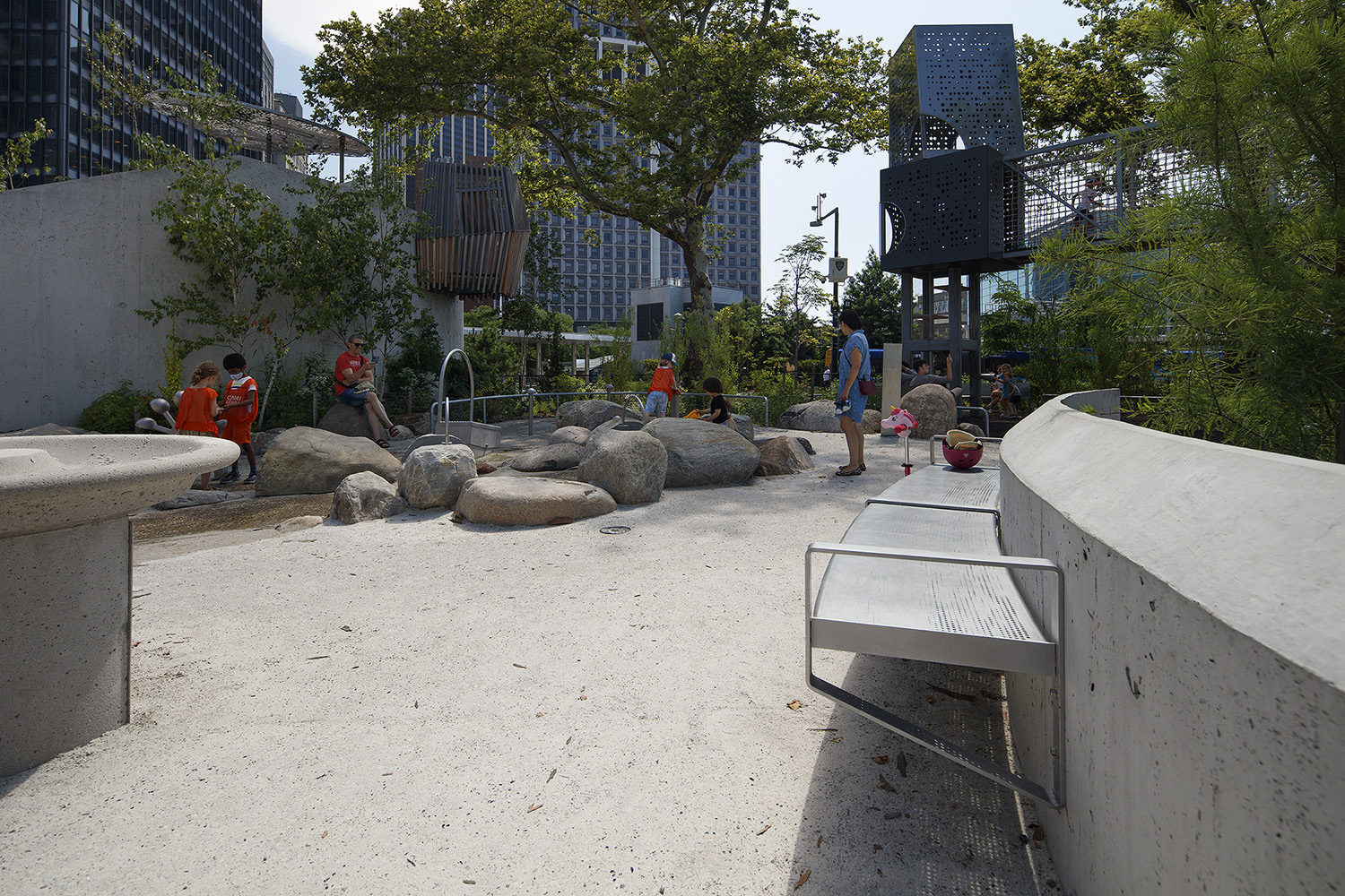 Battery-Playscape_custom-cantilever-bench_002 Children and adults are gathered in a modern urban playground with concrete benches, rocks, play structures, and trees, surrounded by tall buildings.