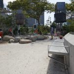 Children and adults are gathered in a modern urban playground with concrete benches, rocks, play structures, and trees, surrounded by tall buildings.
