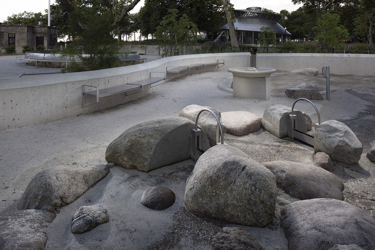 Battery-Playscape_custom-cantilever-bench_003 De gros rochers et des structures de jeux d'eau en métal sont placés dans un espace extérieur en béton avec des bancs incurvés ; des arbres et un bâtiment rond sont visibles à l'arrière-plan.