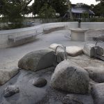 De gros rochers et des structures de jeux d'eau en métal sont placés dans un espace extérieur en béton avec des bancs incurvés ; des arbres et un bâtiment rond sont visibles à l'arrière-plan.