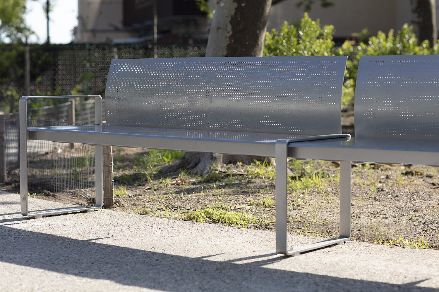 A modern metal bench with a perforated backrest is placed on a sidewalk near a grassy area and trees, with buildings visible in the background.