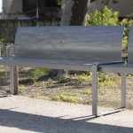 A modern metal bench with a perforated backrest is placed on a sidewalk near a grassy area and trees, with buildings visible in the background.
