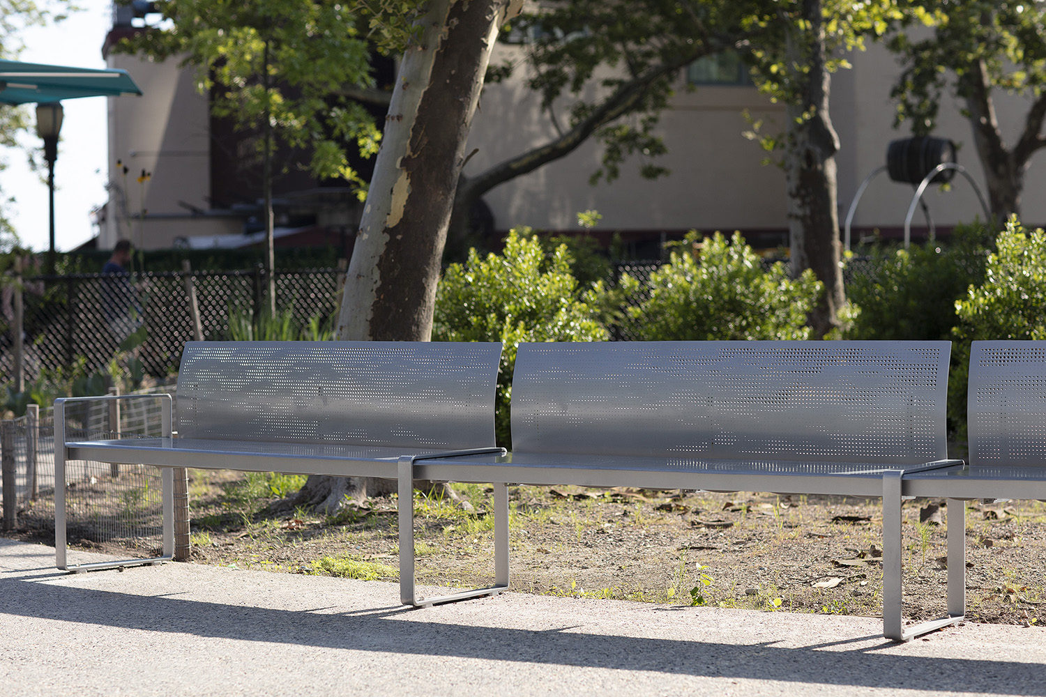 A modern metal bench with perforated backrest and armrests is placed on a paved path beside trees and a fenced area in a park.